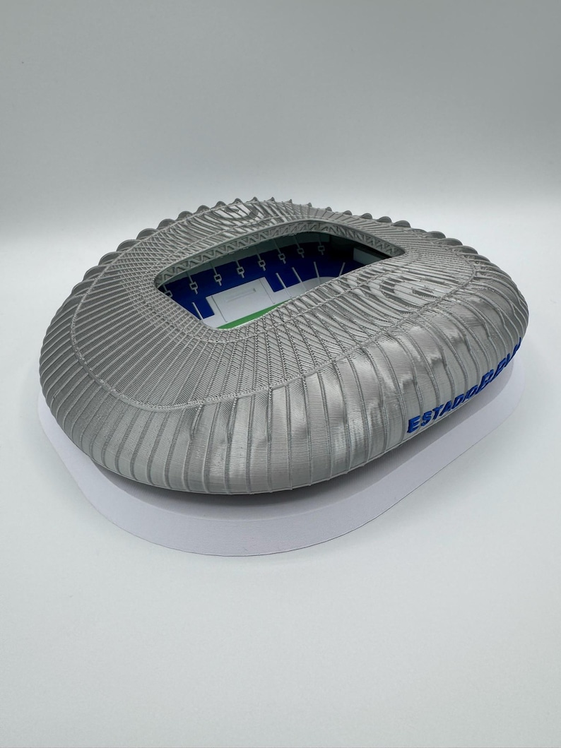 May include: A gray stadium model with a ridged roof and a white base. The interior shows a green field and blue seating. The text "ESTADIO" is visible on the side. A detailed miniature of a sports arena.
