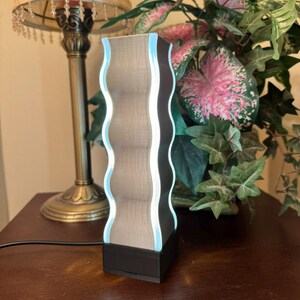 K&ouml;nnte beinhalten: Moderne Lampe mit wellenf&ouml;rmigen Kanten, einem strukturierten, hellbraunen Mittelteil, einem dunkelbraunen Rahmen und einem sanften blauen Leuchten. Die Lampe steht auf einem schwarzen Sockel und ist eingesteckt. Ein dekorativer Artikel f&uuml;r die Inneneinrichtung.