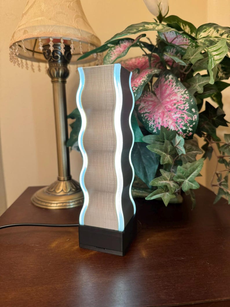 K&ouml;nnte beinhalten: Moderne Lampe mit wellenf&ouml;rmigen Kanten, einem strukturierten, hellbraunen Mittelteil, einem dunkelbraunen Rahmen und einem sanften blauen Leuchten. Die Lampe steht auf einem schwarzen Sockel und ist eingesteckt. Ein dekorativer Artikel f&uuml;r die Inneneinrichtung.