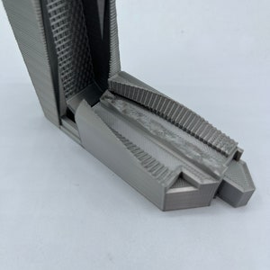 May include: A 3D printed model of a building with a textured surface and a geometric design. The model is made of gray plastic and has a sharp, angular shape.