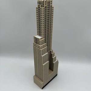 May include: A beige miniature skyscraper model with a detailed facade. The building has a stepped design, with a tall central tower and multiple smaller sections. The model is set against a plain white background.