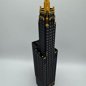 Op de afbeelding: Een gedetailleerd model van een wolkenkrabber met een zwarte gevel en gouden accenten. Het gebouw heeft een gelaagd ontwerp, met een hoge centrale toren en een kortere basis. De top van de toren is versierd met ingewikkelde gouden details.