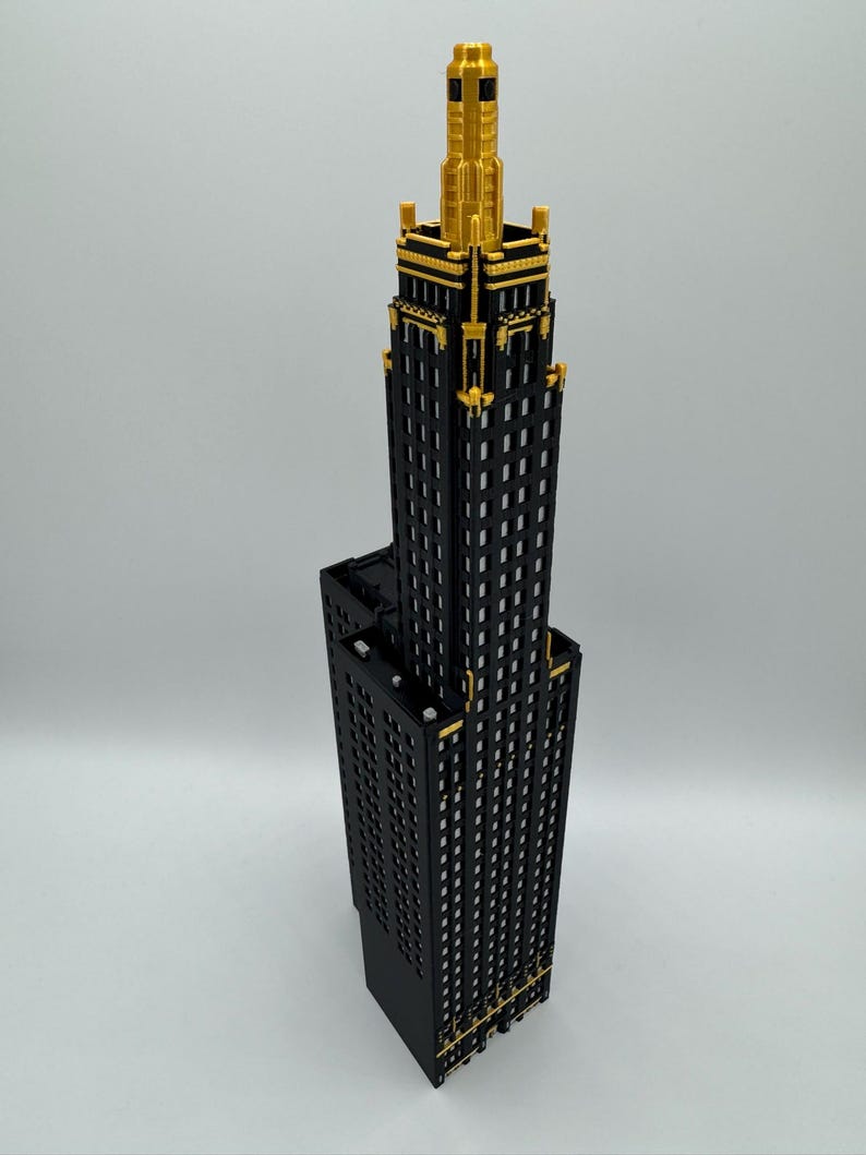 Op de afbeelding: Een gedetailleerd model van een wolkenkrabber met een zwarte gevel en gouden accenten. Het gebouw heeft een gelaagd ontwerp, met een hoge centrale toren en een kortere basis. De top van de toren is versierd met ingewikkelde gouden details.