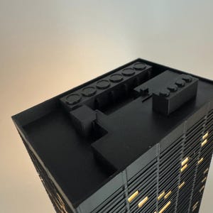 Peut inclure: Maquette architecturale noire d'un gratte-ciel avec des fen&ecirc;tres &eacute;clair&eacute;es. Le mod&egrave;le a un toit plat avec plusieurs &eacute;l&eacute;ments rectangulaires et circulaires. La fa&ccedil;ade du b&acirc;timent pr&eacute;sente des lignes verticales, et les fen&ecirc;tres &eacute;mettent une lumi&egrave;re jaune chaude.
