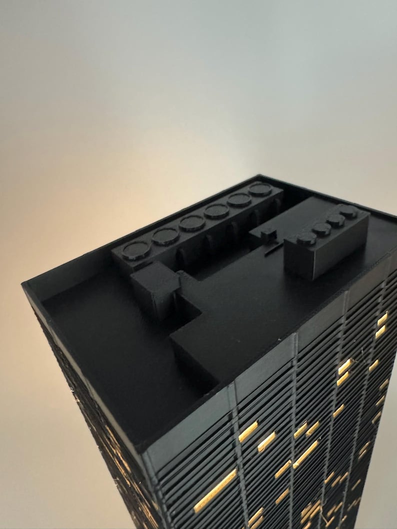 Peut inclure: Maquette architecturale noire d'un gratte-ciel avec des fen&ecirc;tres &eacute;clair&eacute;es. Le mod&egrave;le a un toit plat avec plusieurs &eacute;l&eacute;ments rectangulaires et circulaires. La fa&ccedil;ade du b&acirc;timent pr&eacute;sente des lignes verticales, et les fen&ecirc;tres &eacute;mettent une lumi&egrave;re jaune chaude.