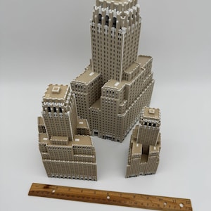 May include: A collection of beige and white miniature building models, including a large skyscraper and several smaller structures. A wooden ruler, approximately 30 cm long, is placed near the buildings for scale.