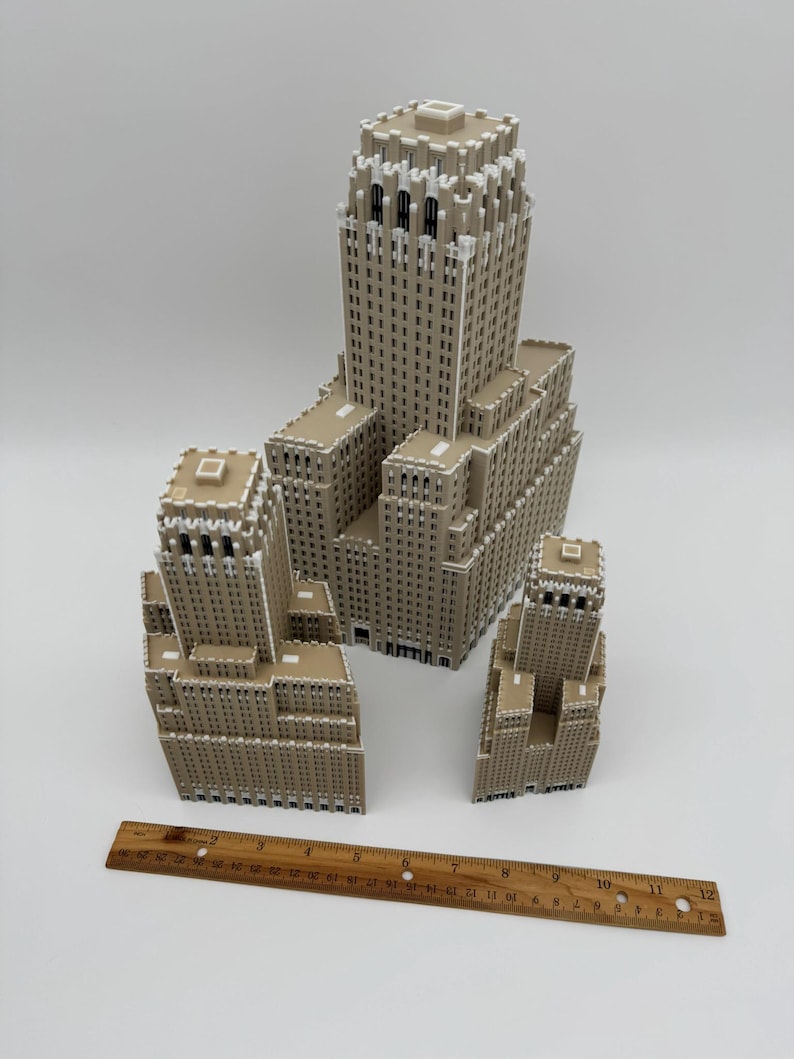 May include: A collection of beige and white miniature building models, including a large skyscraper and several smaller structures. A wooden ruler, approximately 30 cm long, is placed near the buildings for scale.