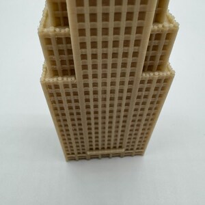May include: A tan-colored, miniature replica of a skyscraper, featuring a detailed grid pattern across its facade. The model has multiple tiers, with a textured surface, and is likely a decorative item or architectural model.