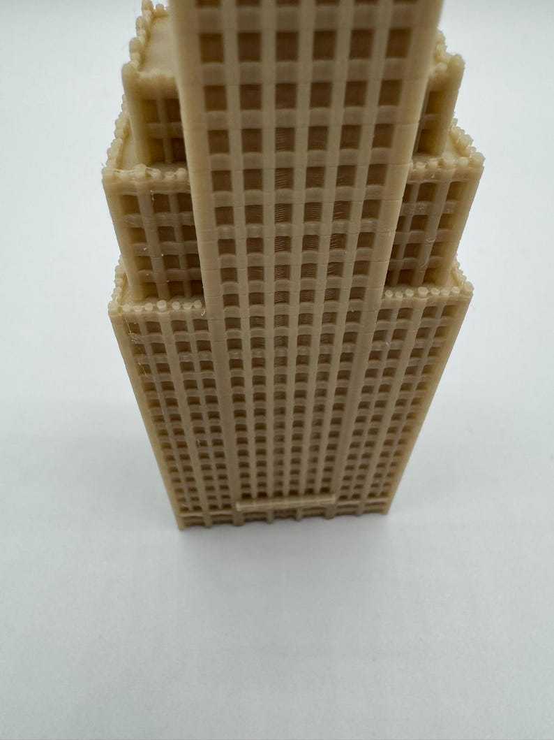 May include: A tan-colored, miniature replica of a skyscraper, featuring a detailed grid pattern across its facade. The model has multiple tiers, with a textured surface, and is likely a decorative item or architectural model.