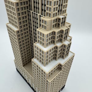 May include: A detailed 3D-printed model of a skyscraper, featuring multiple tiers and a complex architectural design. The building is primarily beige with white accents, showcasing a realistic representation of a modern high-rise structure. The model sits on a black base.