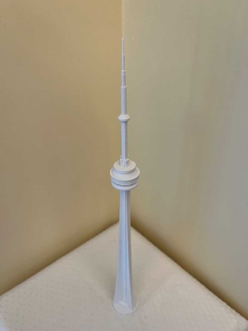 May include: A white miniature of the CN Tower, a tall structure with a wide base and a slender, spire-like top. The model is made of a solid, opaque material and stands on a white surface. The tower's design includes a circular observation deck.
