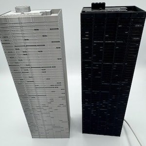 Peut inclure: Deux maquettes de gratte-ciel miniatures. L'une est gris clair, l'autre est noire. Les deux ont une forme rectangulaire avec des lignes horizontales repr&eacute;sentant des fen&ecirc;tres. Les b&acirc;timents sont sur une surface blanche.