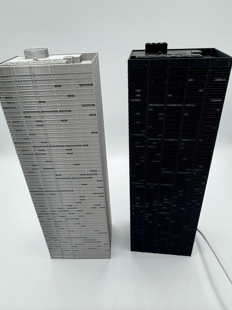 Peut inclure: Deux maquettes de gratte-ciel miniatures. L'une est gris clair, l'autre est noire. Les deux ont une forme rectangulaire avec des lignes horizontales repr&eacute;sentant des fen&ecirc;tres. Les b&acirc;timents sont sur une surface blanche.