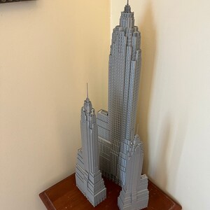 K&ouml;nnte beinhalten: Ein graues Modell des Empire State Building und anderer Wolkenkratzer. Die Geb&auml;ude stehen auf einem kleinen Holztisch. Ein durchsichtiges Lineal liegt vor den Geb&auml;uden. Die Geb&auml;ude sind grau und detailliert.