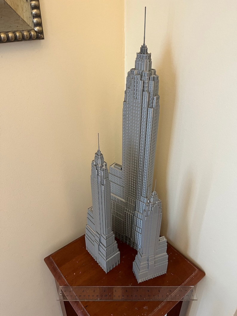 K&ouml;nnte beinhalten: Ein graues Modell des Empire State Building und anderer Wolkenkratzer. Die Geb&auml;ude stehen auf einem kleinen Holztisch. Ein durchsichtiges Lineal liegt vor den Geb&auml;uden. Die Geb&auml;ude sind grau und detailliert.