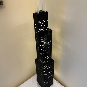 May include: A black model of the Willis Tower, formerly known as the Sears Tower, with white antenna details. The model is lit up with white lights, creating a realistic nighttime cityscape effect.