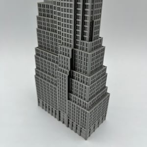 K&ouml;nnte beinhalten: Eine graue Miniatur des Empire State Building. Das Modell hat ein abgestuftes Design mit zahlreichen Fenstern und architektonischen Details. Das Geb&auml;ude ist einfarbig grau und steht auf einer wei&szlig;en Oberfl&auml;che.