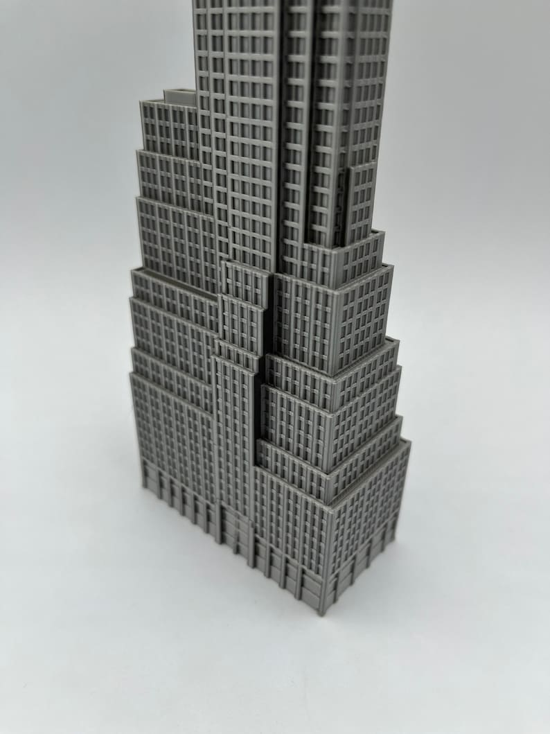 K&ouml;nnte beinhalten: Eine graue Miniatur des Empire State Building. Das Modell hat ein abgestuftes Design mit zahlreichen Fenstern und architektonischen Details. Das Geb&auml;ude ist einfarbig grau und steht auf einer wei&szlig;en Oberfl&auml;che.