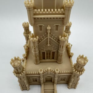 May include: A beige miniature castle model with multiple turrets and architectural details. The model features a central tower and several smaller towers, with detailed windows and crenellations. The overall design is intricate and symmetrical, showcasing a detailed miniature building.