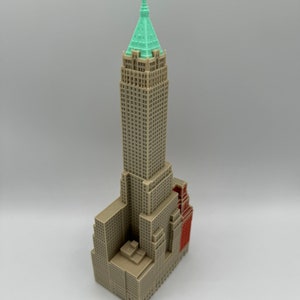 May include: A miniature model of a tall, brown building with a green roof and a red accent. The building has many windows and a detailed facade.