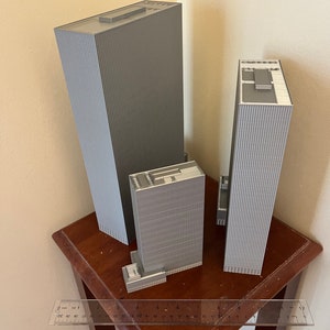 May include: Three gray 3D printed model buildings of varying sizes on a wooden table with a ruler for scale. The tallest building is 12 inches tall.