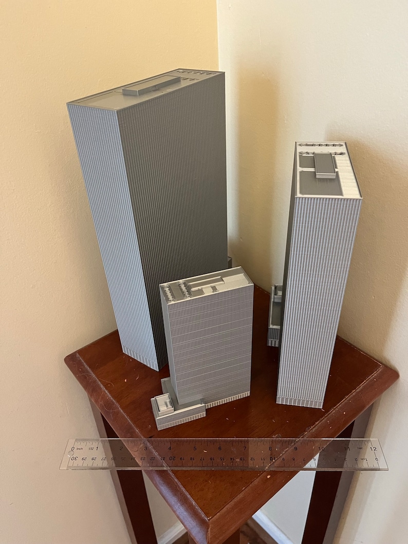 May include: Three gray 3D printed model buildings of varying sizes on a wooden table with a ruler for scale. The tallest building is 12 inches tall.