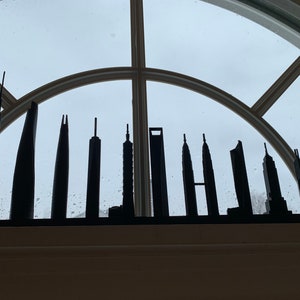 May include: A black skyline silhouette featuring various skyscrapers. The architectural models are displayed on a windowsill against a cloudy sky. The buildings vary in height and design, creating a cityscape.