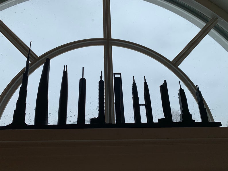May include: A black skyline silhouette featuring various skyscrapers. The architectural models are displayed on a windowsill against a cloudy sky. The buildings vary in height and design, creating a cityscape.