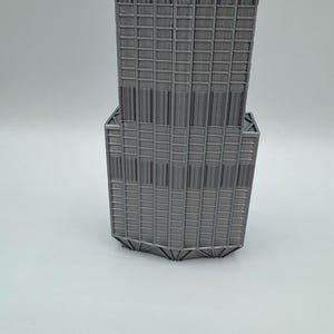 May include: A gray 3D printed model of a skyscraper featuring a stepped architectural design with vertical columns and a grid of rectangular windows. The structure exhibits a distinct tiered silhouette with a wider base section and a narrower upper tower.