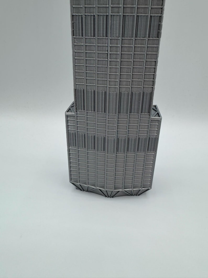 May include: A gray 3D printed model of a skyscraper featuring a stepped architectural design with vertical columns and a grid of rectangular windows. The structure exhibits a distinct tiered silhouette with a wider base section and a narrower upper tower.