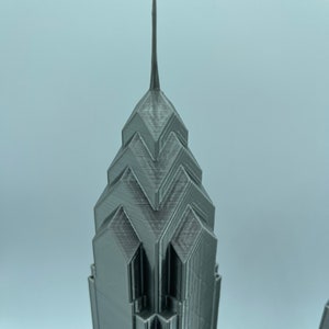 May include: A 3D printed model of a skyscraper with a pointed top and a geometric design. The model is made of a gray plastic material.