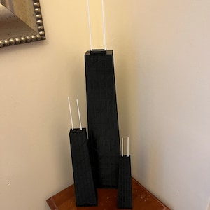 May include: Three black architectural models of a skyscraper, each with two white antennae. The models are of varying sizes, with the largest in the center. The models are on a small wooden table.