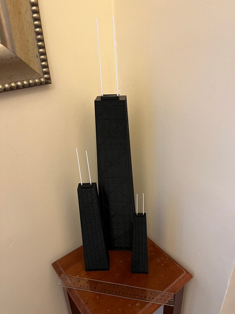 May include: Three black architectural models of a skyscraper, each with two white antennae. The models are of varying sizes, with the largest in the center. The models are on a small wooden table.