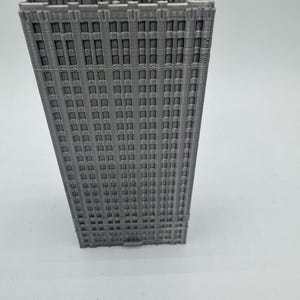 May include: A grey 3D printed model of a tall building with many windows. The building has a simple design with a flat roof and a rectangular shape.