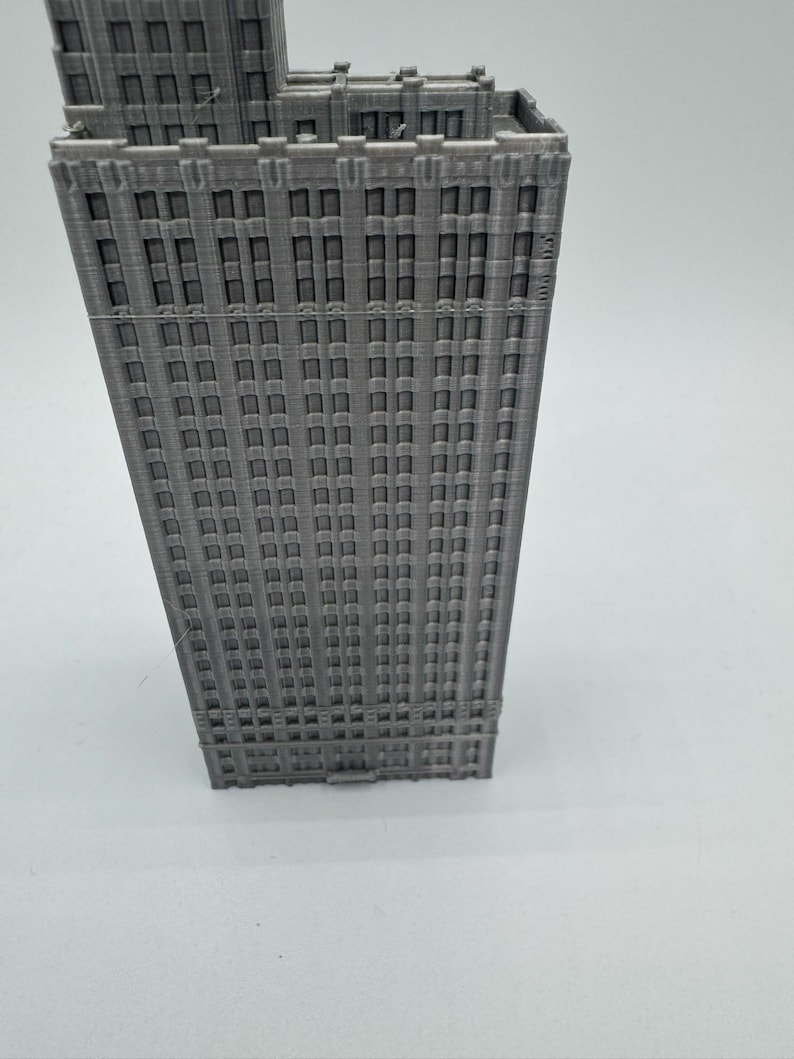 May include: A grey 3D printed model of a tall building with many windows. The building has a simple design with a flat roof and a rectangular shape.