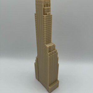 May include: A beige-colored miniature skyscraper model with a detailed facade. The building has a rectangular base and a tall, tiered structure with numerous windows. The model is a representation of a city building.