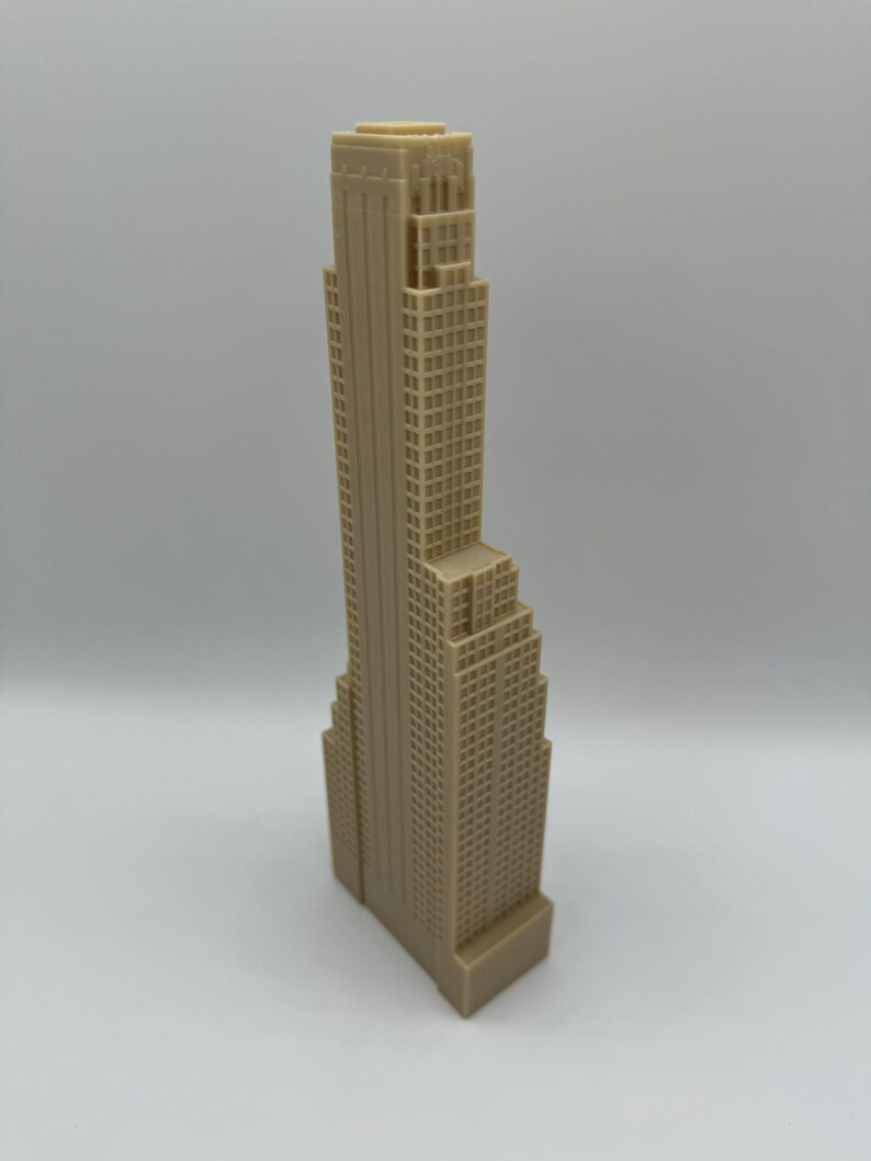 May include: A beige-colored miniature skyscraper model with a detailed facade. The building has a rectangular base and a tall, tiered structure with numerous windows. The model is a representation of a city building.