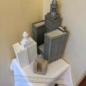 May include: Four miniature building models, two are white and two are gray. The tallest building is gray and has a pointed roof. The models are arranged on a white tablecloth with a ruler in the foreground.
