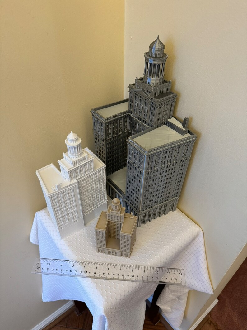 May include: Four miniature building models, two are white and two are gray. The tallest building is gray and has a pointed roof. The models are arranged on a white tablecloth with a ruler in the foreground.