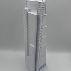 May include: A light gray architectural model of a tall skyscraper. The model has a rectangular shape with detailed exterior features, including vertical lines, geometric patterns, and a complex top structure. The building is a modern design.