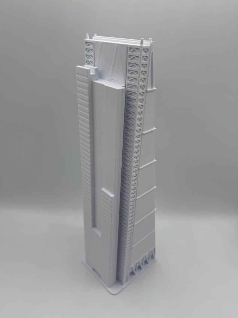 May include: A light gray architectural model of a tall skyscraper. The model has a rectangular shape with detailed exterior features, including vertical lines, geometric patterns, and a complex top structure. The building is a modern design.