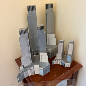 Op de afbeelding: Een set van vijf 3D-geprinte grijze wolkenkrabbers, elk met een uniek ontwerp, gerangschikt op een houten tafel met een liniaal voor schaal.