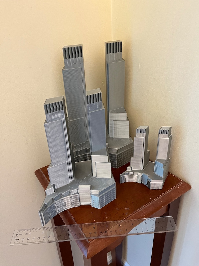 Op de afbeelding: Een set van vijf 3D-geprinte grijze wolkenkrabbers, elk met een uniek ontwerp, gerangschikt op een houten tafel met een liniaal voor schaal.