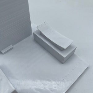 K&ouml;nnte beinhalten: Wei&szlig;es 3D-gedrucktes Modell eines Geb&auml;udes mit Flachdach und einem kleinen rechteckigen Anbau an der Seite. Das Geb&auml;ude steht auf einem wei&szlig;en Sockel.