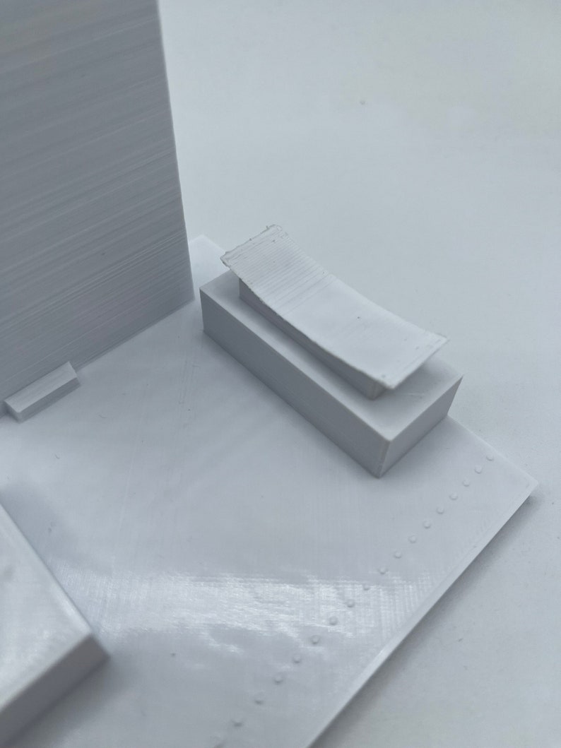 K&ouml;nnte beinhalten: Wei&szlig;es 3D-gedrucktes Modell eines Geb&auml;udes mit Flachdach und einem kleinen rechteckigen Anbau an der Seite. Das Geb&auml;ude steht auf einem wei&szlig;en Sockel.