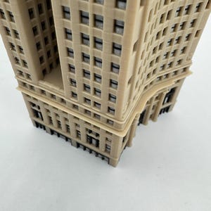 Op de afbeelding: Een beige miniatuurmodel van een hoog gebouw met een gebogen gevel en veel ramen. Het model is gemaakt van plastic en heeft een gedetailleerd ontwerp.