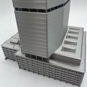 Puede incluir: Un modelo de rascacielos moderno impreso en 3D con un acabado gris. El edificio tiene una base rectangular y una torre alta y esbelta con una serie de ventanas y retrocesos.