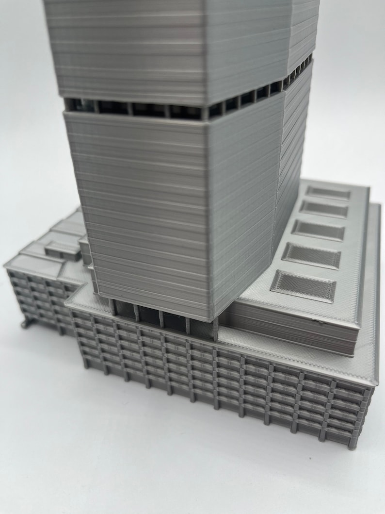 Puede incluir: Un modelo de rascacielos moderno impreso en 3D con un acabado gris. El edificio tiene una base rectangular y una torre alta y esbelta con una serie de ventanas y retrocesos.