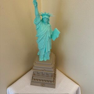 May include: A teal green statue of the Statue of Liberty with a brown base. The statue is holding a book in her left hand and a torch in her right hand.