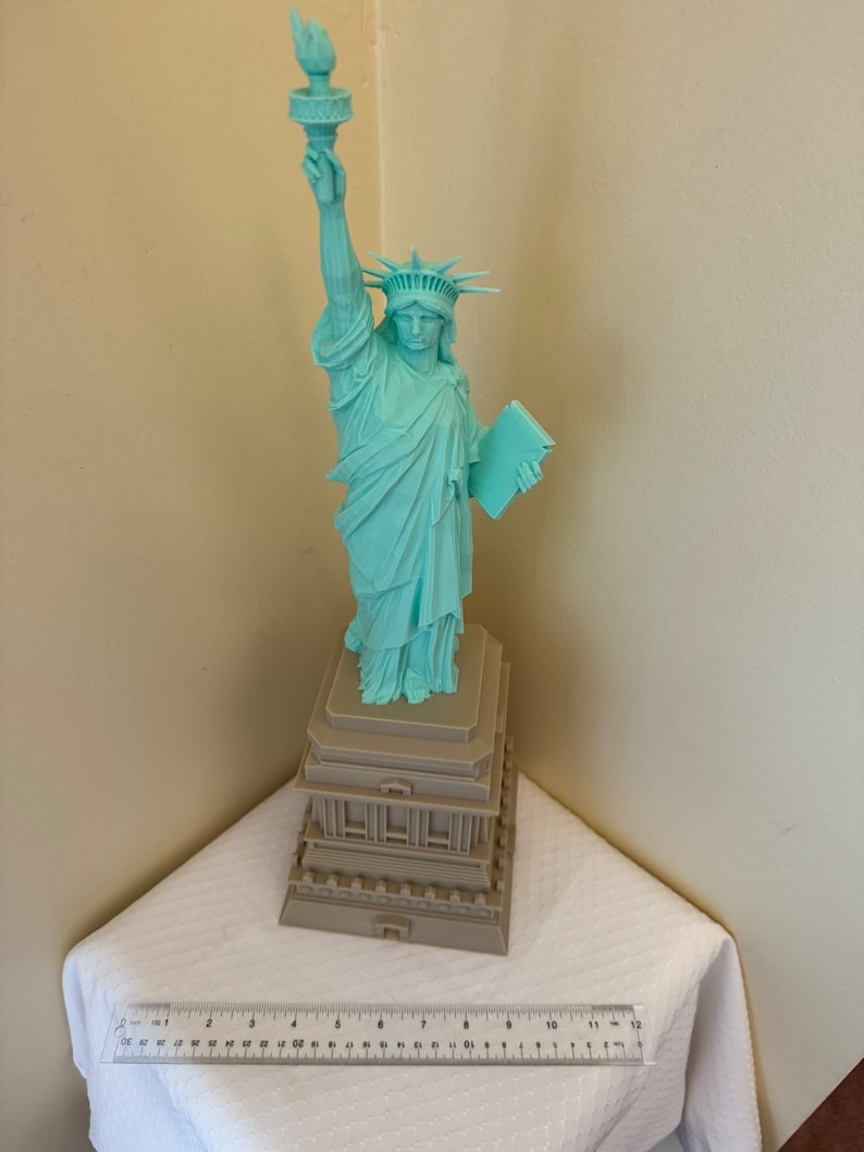 May include: A teal green statue of the Statue of Liberty with a brown base. The statue is holding a book in her left hand and a torch in her right hand.